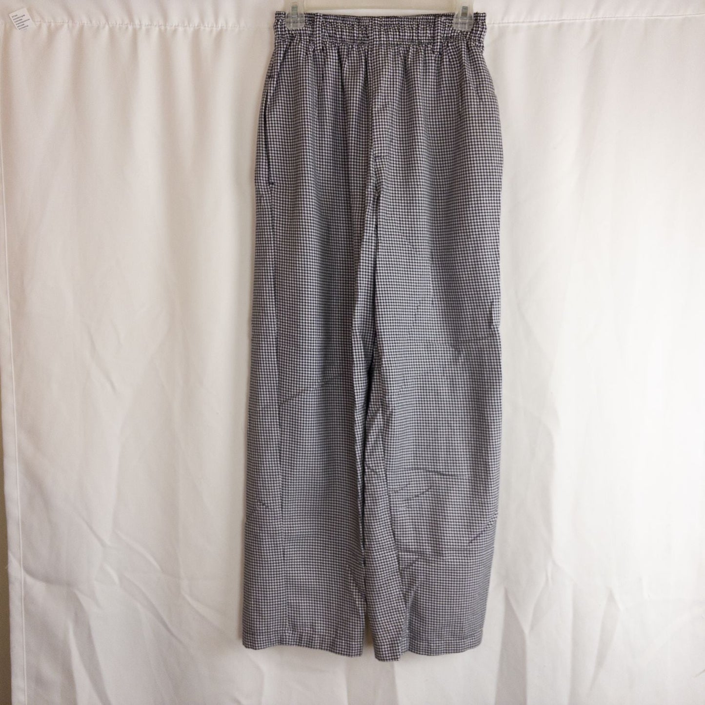 Mercer Culinary black and white houndstooth high rise pants in womens small size hanging on plastic hanger against white background showing front, side pockets, and back Mercer Culinary logo