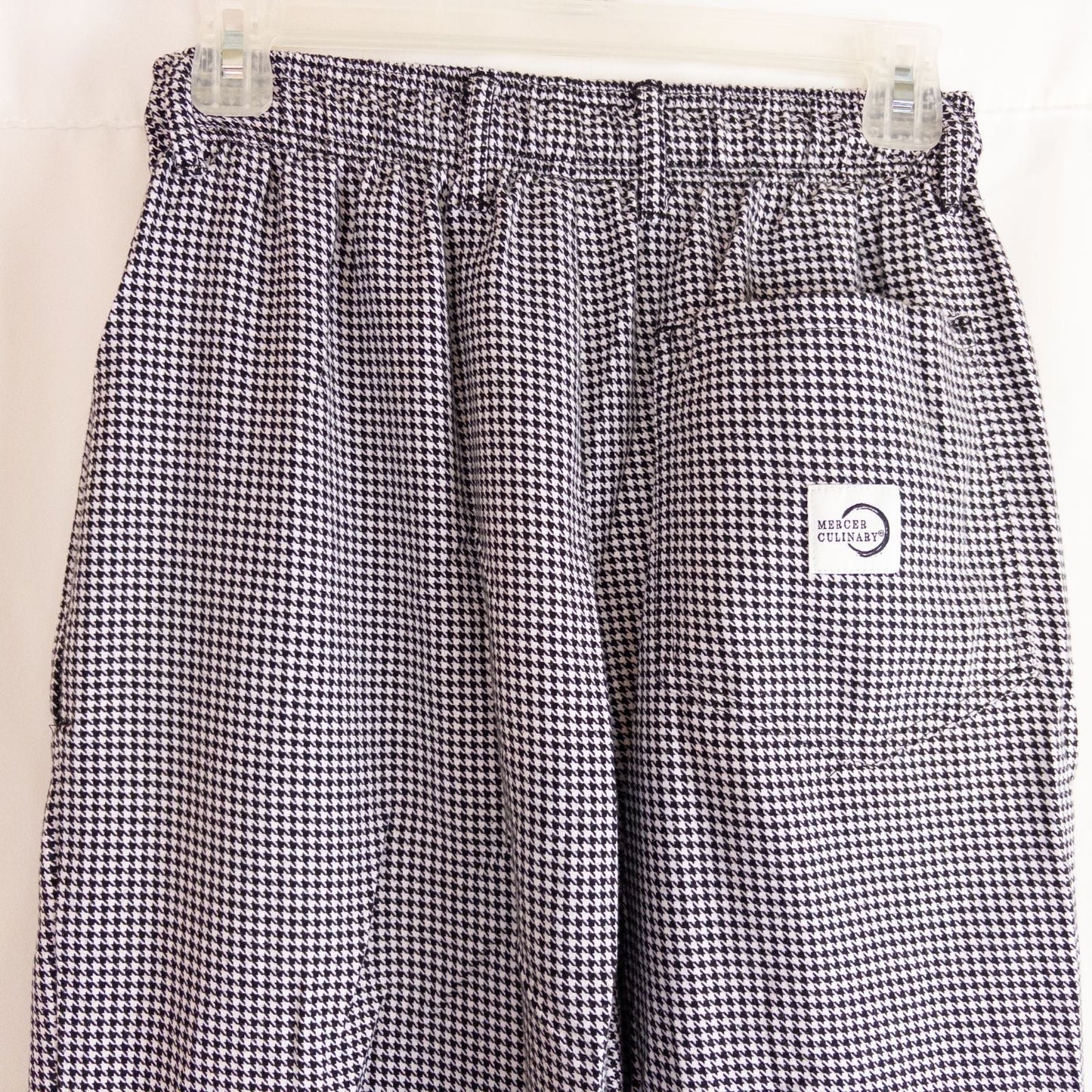 Mercer Culinary black and white houndstooth high rise pants in womens small size hanging on plastic hanger against white background showing front, side pockets, and back Mercer Culinary logo image 6