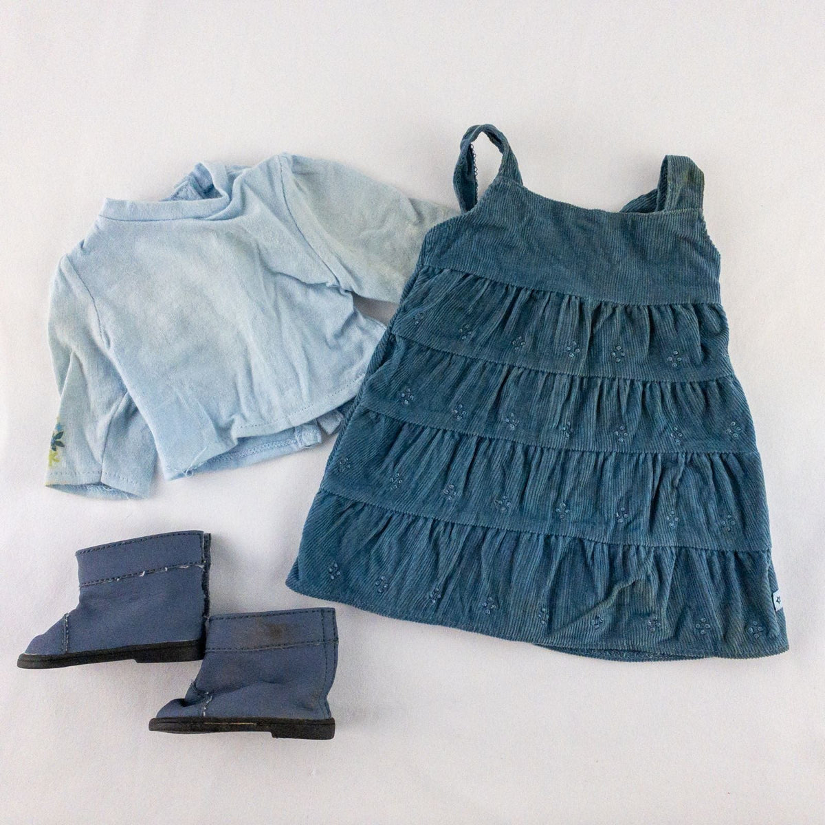 American Girl doll jumper dress outfit with blue corduroy jumper dress, faded light blue long sleeve top, and blue doll boots laid flat on white background