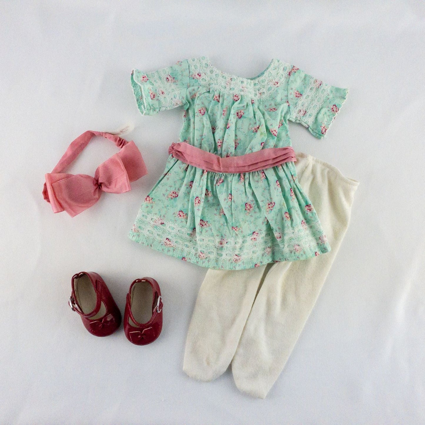 American Girl doll dress set including green floral doll dress with lace trim and pink bow sash, pink bow headband, white stockings, and red patent shoes