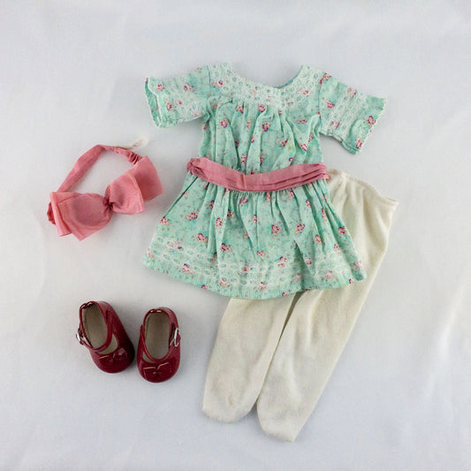 American Girl doll dress set including green floral doll dress with lace trim and pink bow sash, pink bow headband, white stockings, and red patent shoes