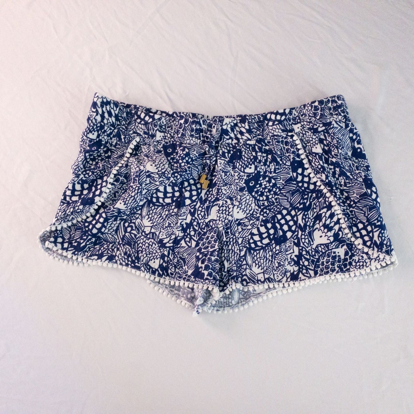 Lily Pullitzer blue fish print lounge shorts with pom pom trim and drawstring waistband laid flat