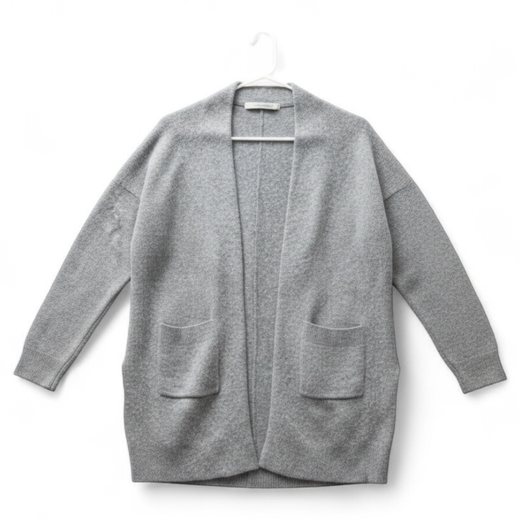 Athleta gray wool cashmere open knit sweater cardigan hanging on white door with two front pockets and ribbed cuffs, showing small brown speck on pocket and fuzzy texture front on white muslin photography backdrop